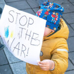 A young child holds up a sign reading "Stop the war!" with the colours of the Ukraine flag.