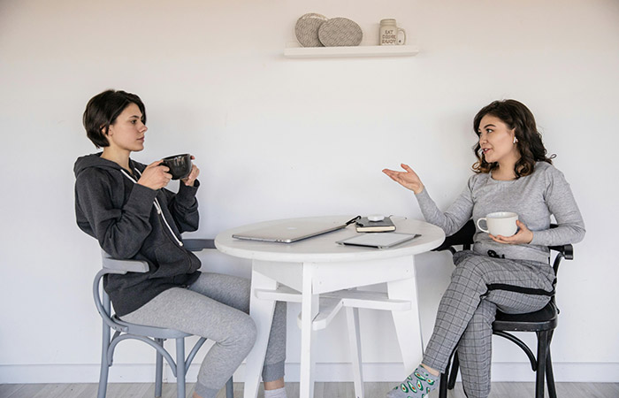 Two women sit at a table drinking coffee or tea and having a discussion